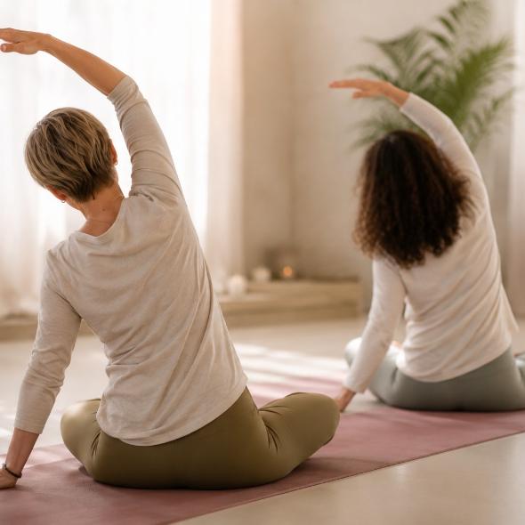 Zwei Frauen üben im Sitzen eine seitliche Dehnung auf der Yogamatte